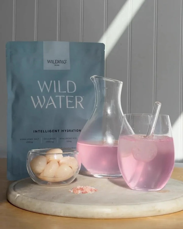 WILD WATER™ pouch beside a carafe and glass of pink lychee drink, with lychees and pink salt